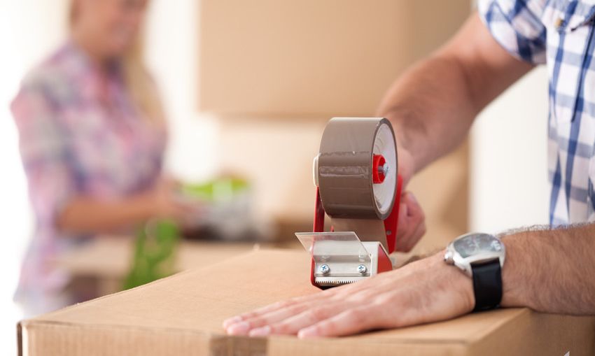 Man in a checkered shirt taping a moving box closed.